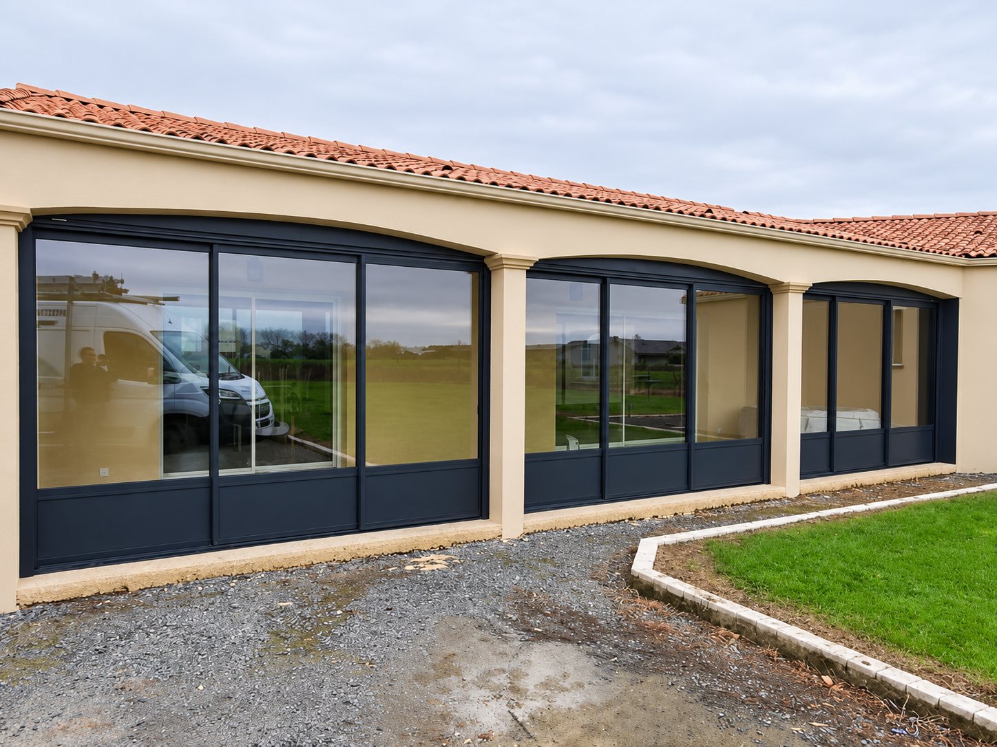 Façade d'une maison de plain-pied à toit de tuiles équipée de grandes baies vitrées coulissantes anthracite sous des arcades, avec une allée de gravier et une pelouse au premier plan.