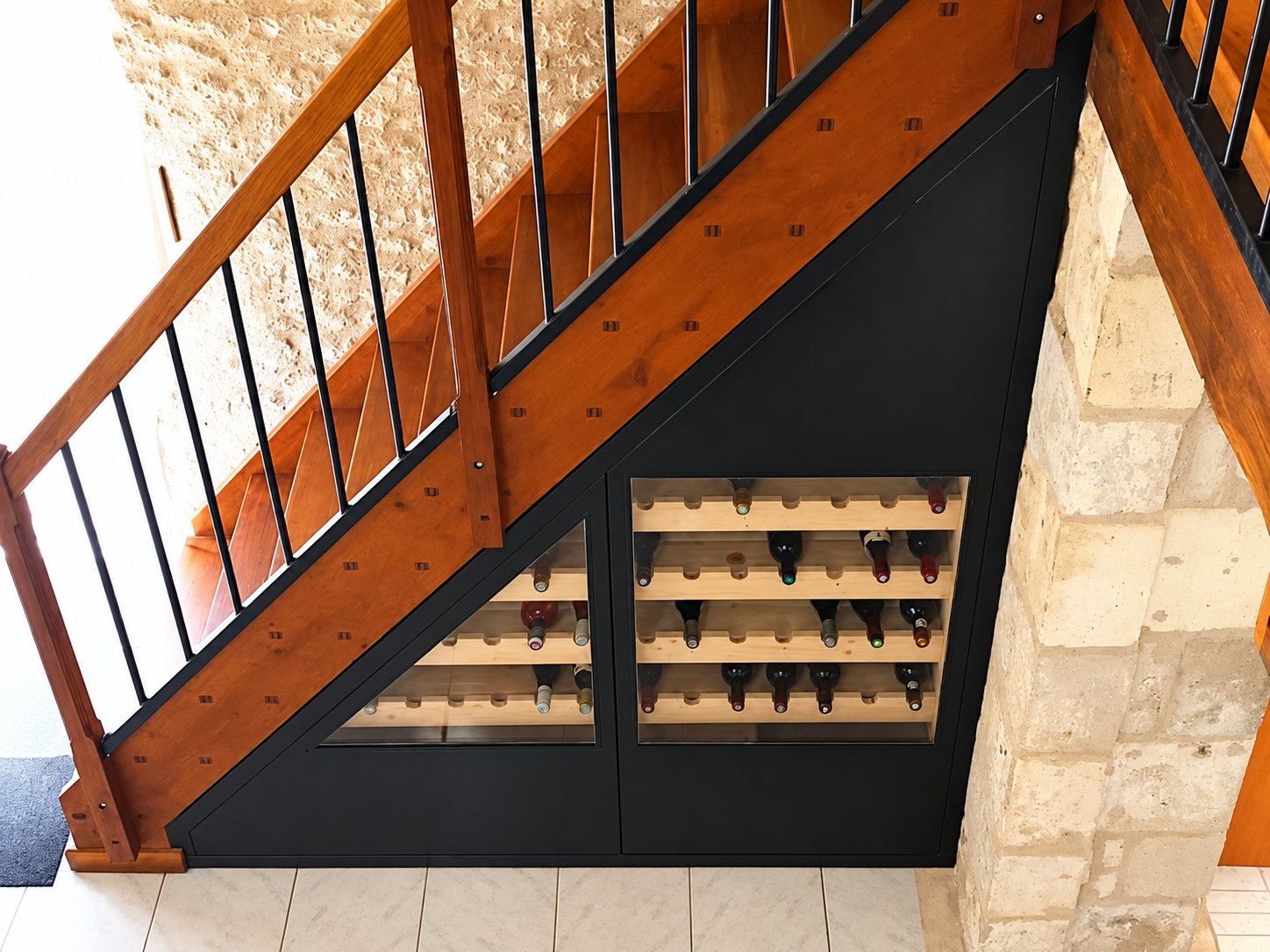 Un casier à bouteilles encastré derrière des vitres sous un escalier en bois, à côté d'un mur en pierre.