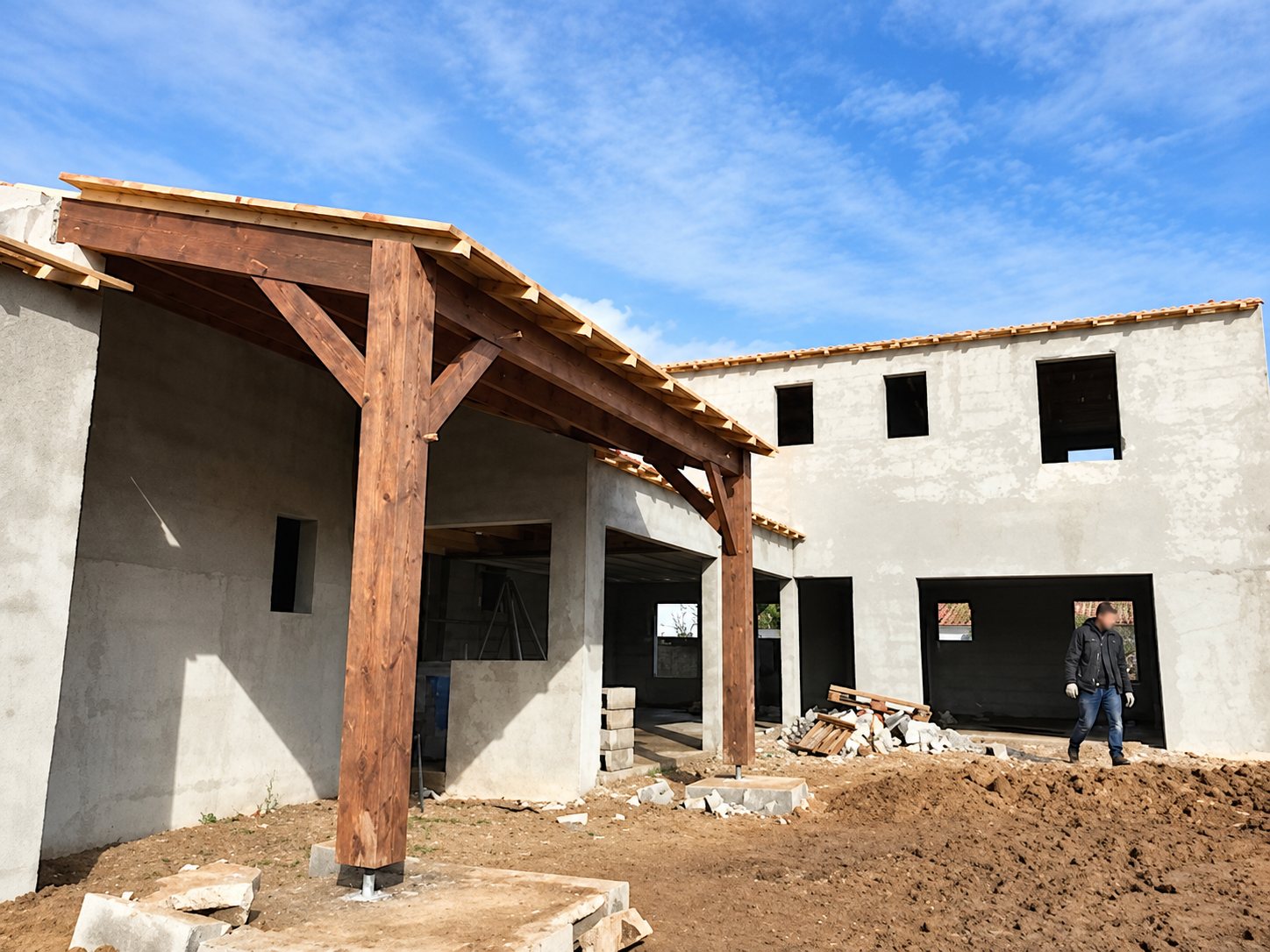 Un chantier de construction d'une maison aux murs bruts en béton, avec une terrasse couverte par une charpente en bois et un homme debout à droite sur la terre.