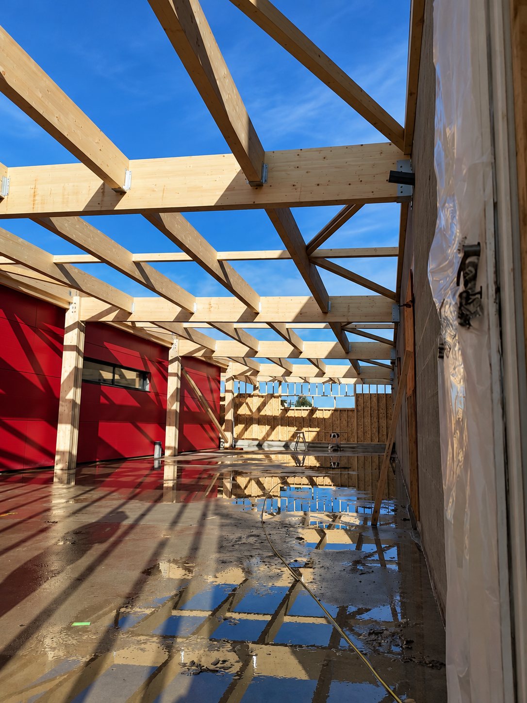 Structure de charpente en bois sans toiture sur un bâtiment aux murs rouges, avec un sol mouillé reflétant le ciel.