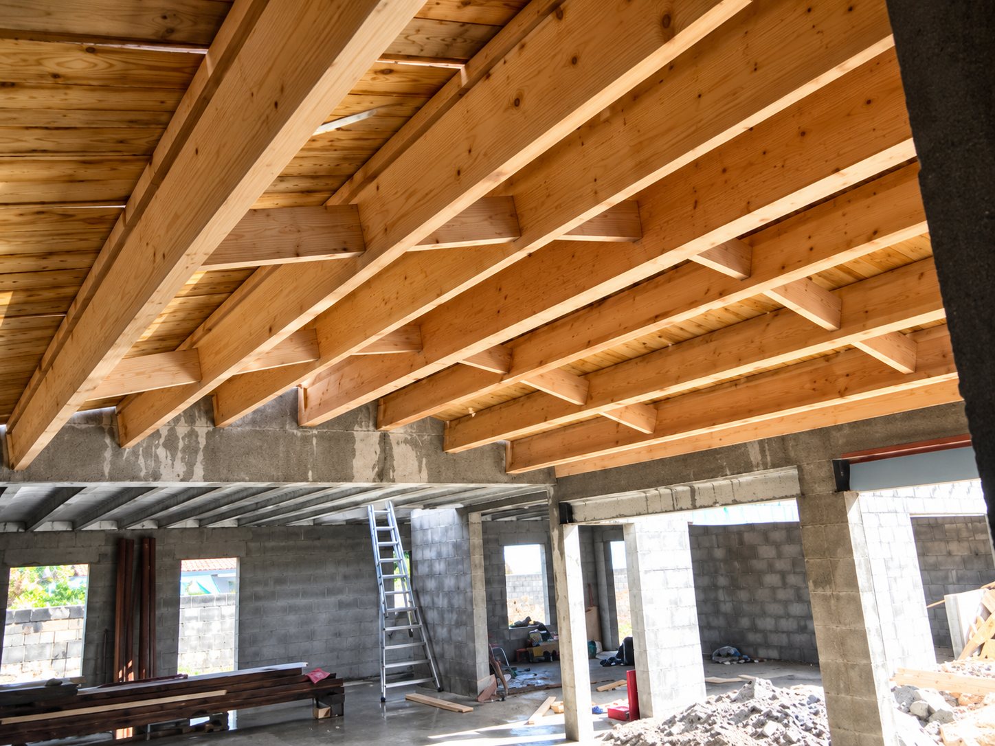 Vue de l'intérieur d'un bâtiment en chantier avec une charpente en bois clair au plafond, des murs en parpaings et une échelle au fond.