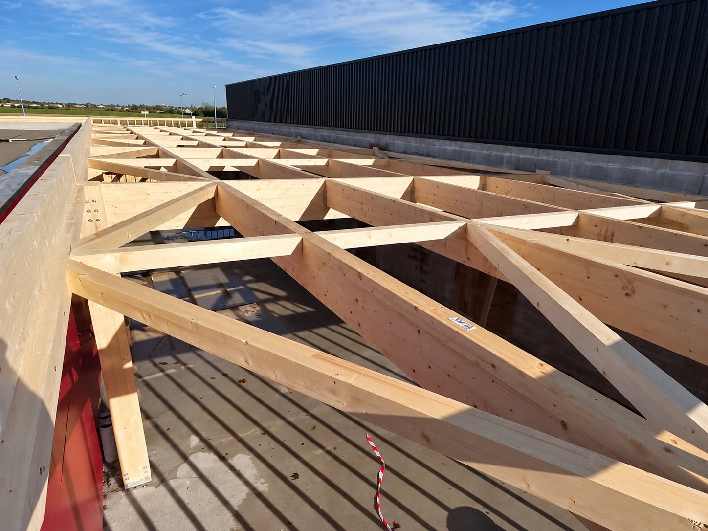 Des fermes de charpente en bois clair sont posées sur la toiture d'un bâtiment à bardage sombre, sous un ciel bleu.