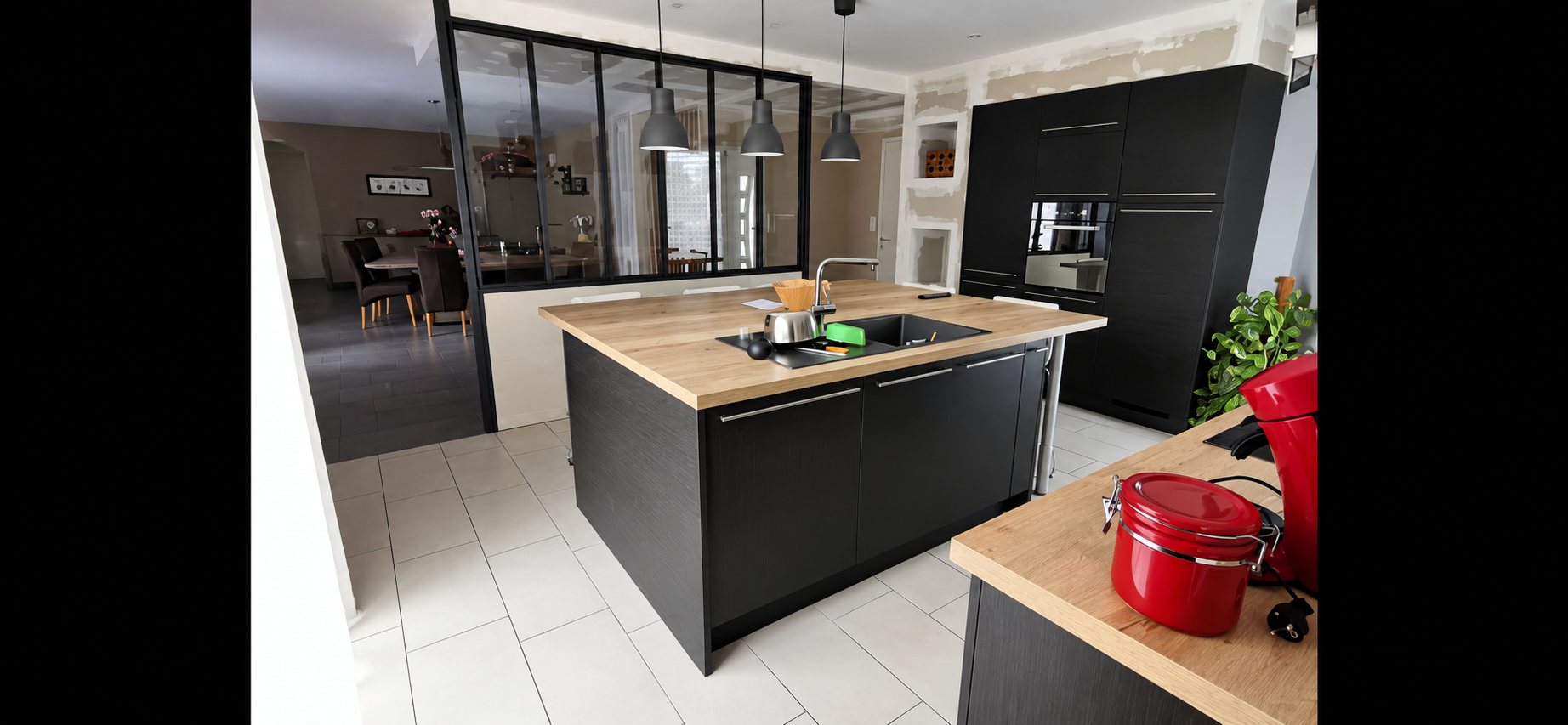 Cuisine contemporaine aux meubles noirs avec un îlot central à plan de travail en bois, une verrière et un sol en carrelage blanc.