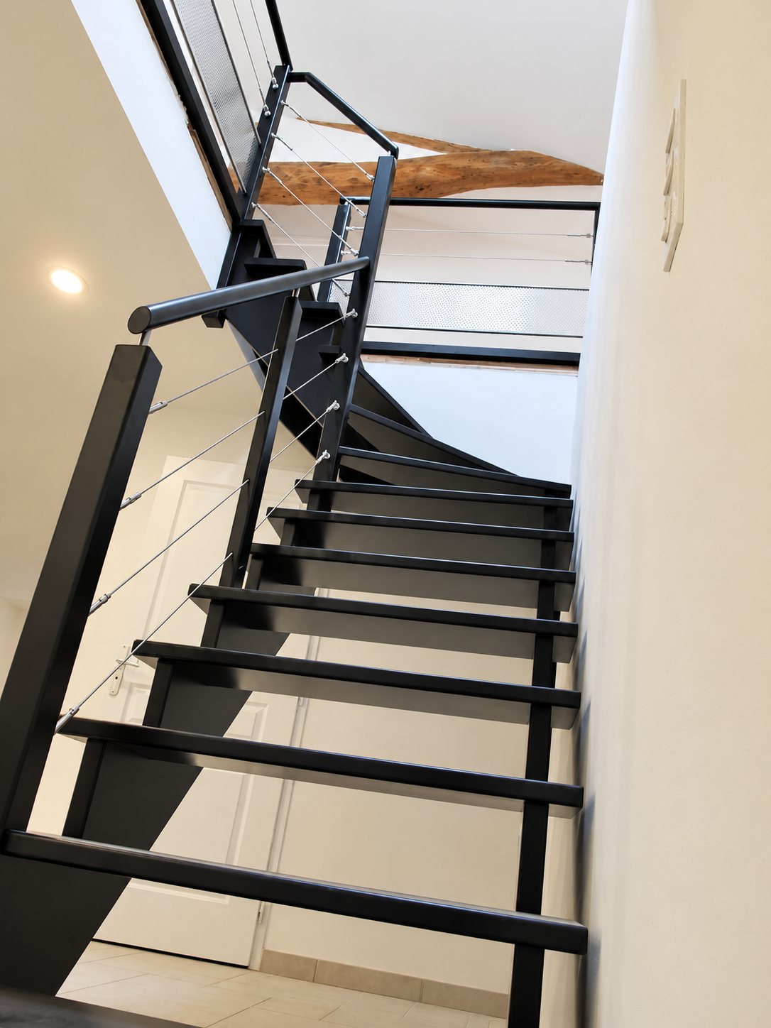 Vue en contre-plongée d'un escalier intérieur noir à quart tournant, avec marches ouvertes, main courante métallique et câbles en inox, près d'une poutre en bois apparente.