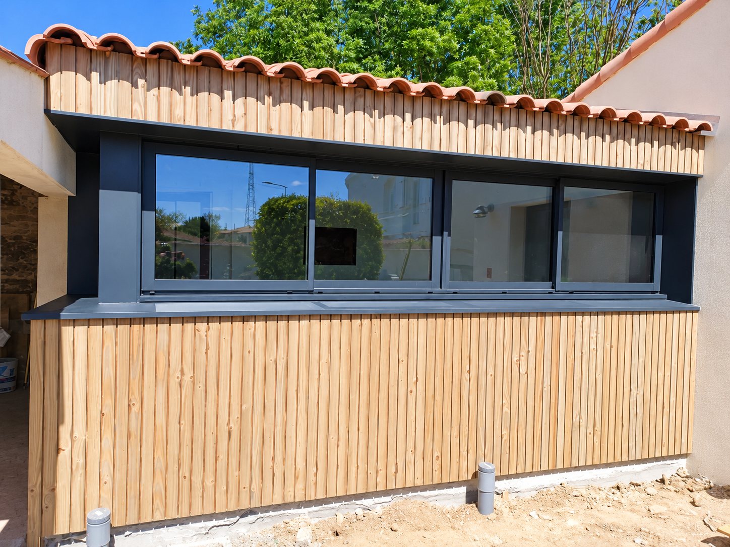 Façade d'une extension recouverte de bardage bois vertical, équipée d'une grande baie vitrée coulissante anthracite sous un toit en tuiles.
