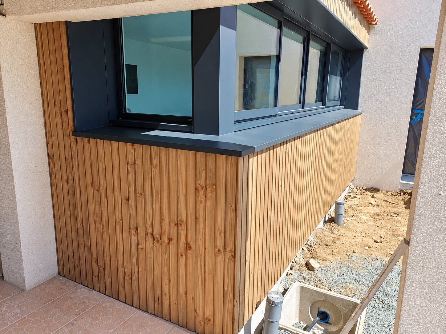 Angle d'une extension de maison recouverte d'un bardage en lames de bois verticales, surmontée de fenêtres coulissantes à encadrement gris anthracite, avec un chantier de terrassement au premier plan.