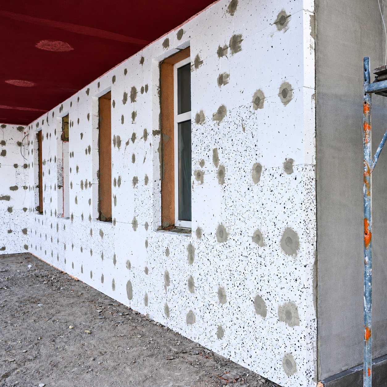 Mur extérieur d'un bâtiment en chantier recouvert de panneaux isolants en polystyrène blanc fixés par des chevilles, avec des fenêtres et un échafaudage à droite.