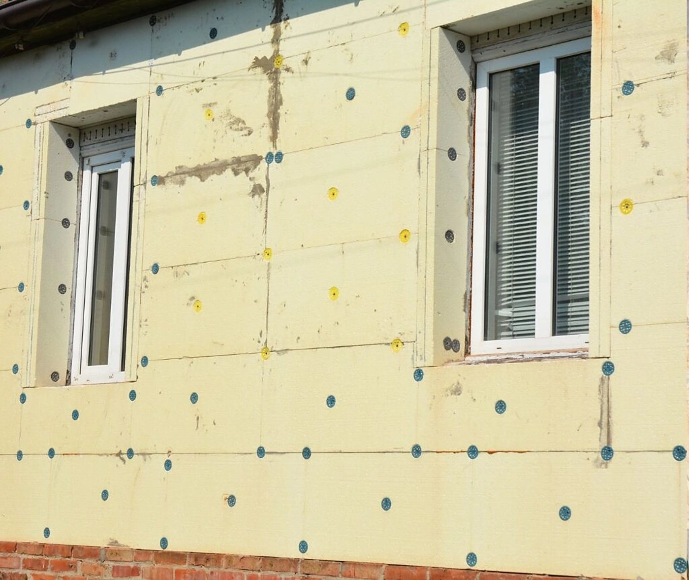 Façade d'une maison dont les panneaux isolants jaunes, fixés par des chevilles bleues et jaunes, entourent deux fenêtres blanches, au-dessus d'un soubassement en briques.