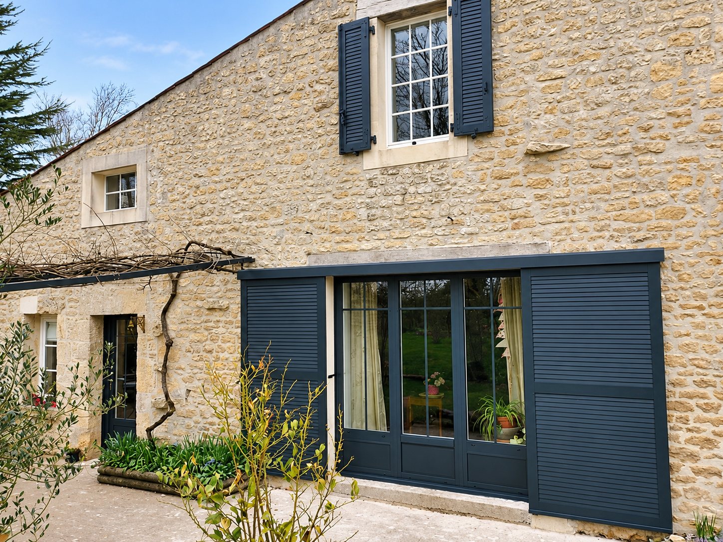 Façade d'une maison en pierre ancienne équipée d'une grande baie vitrée à petits carreaux et de volets coulissants à lames gris anthracite, avec des volets battants à la fenêtre de l'étage.