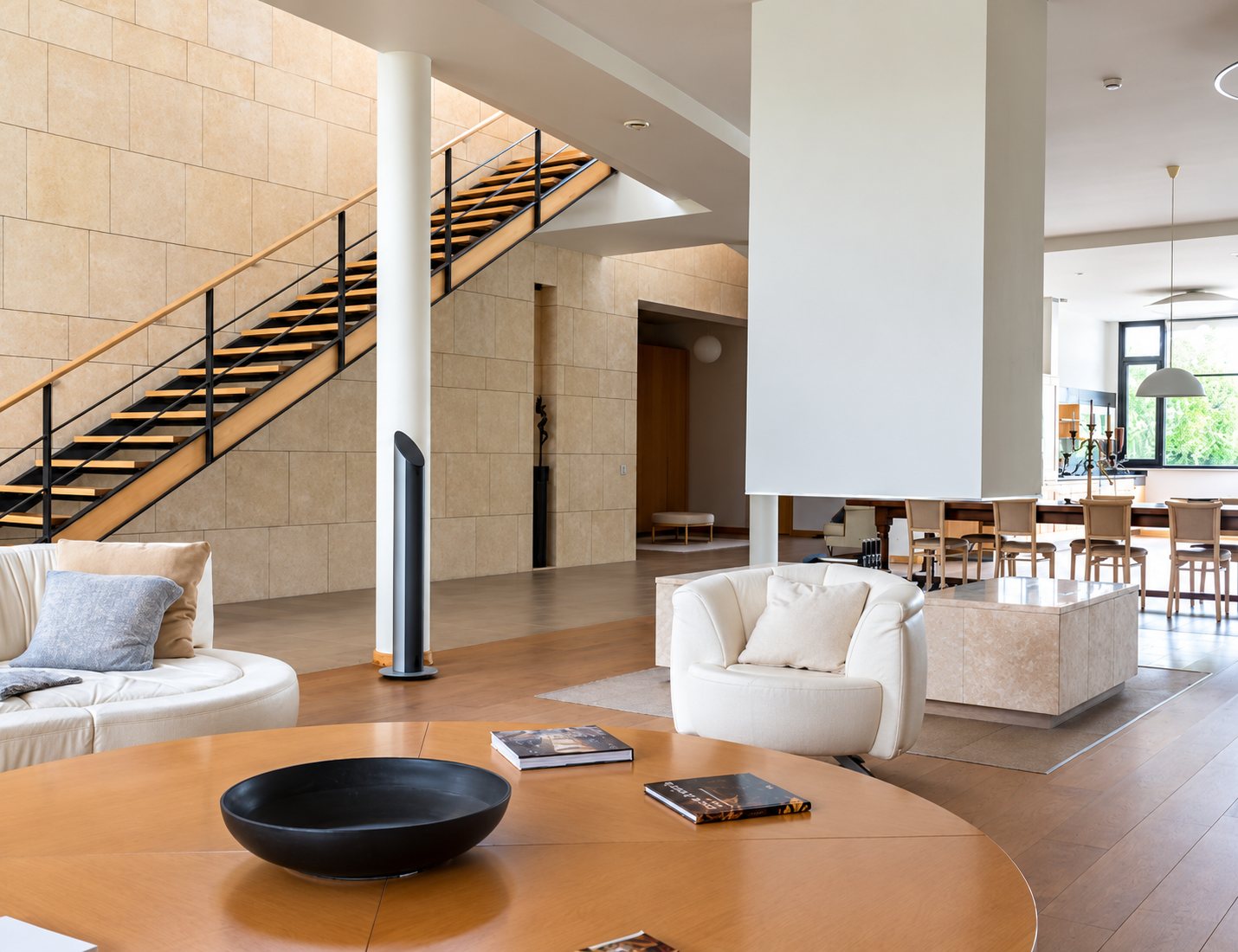 Vaste salon moderne aux murs de pierre claire, avec escalier à marches en bois, fauteuils blancs, table ronde en bois et salle à manger en arrière-plan.