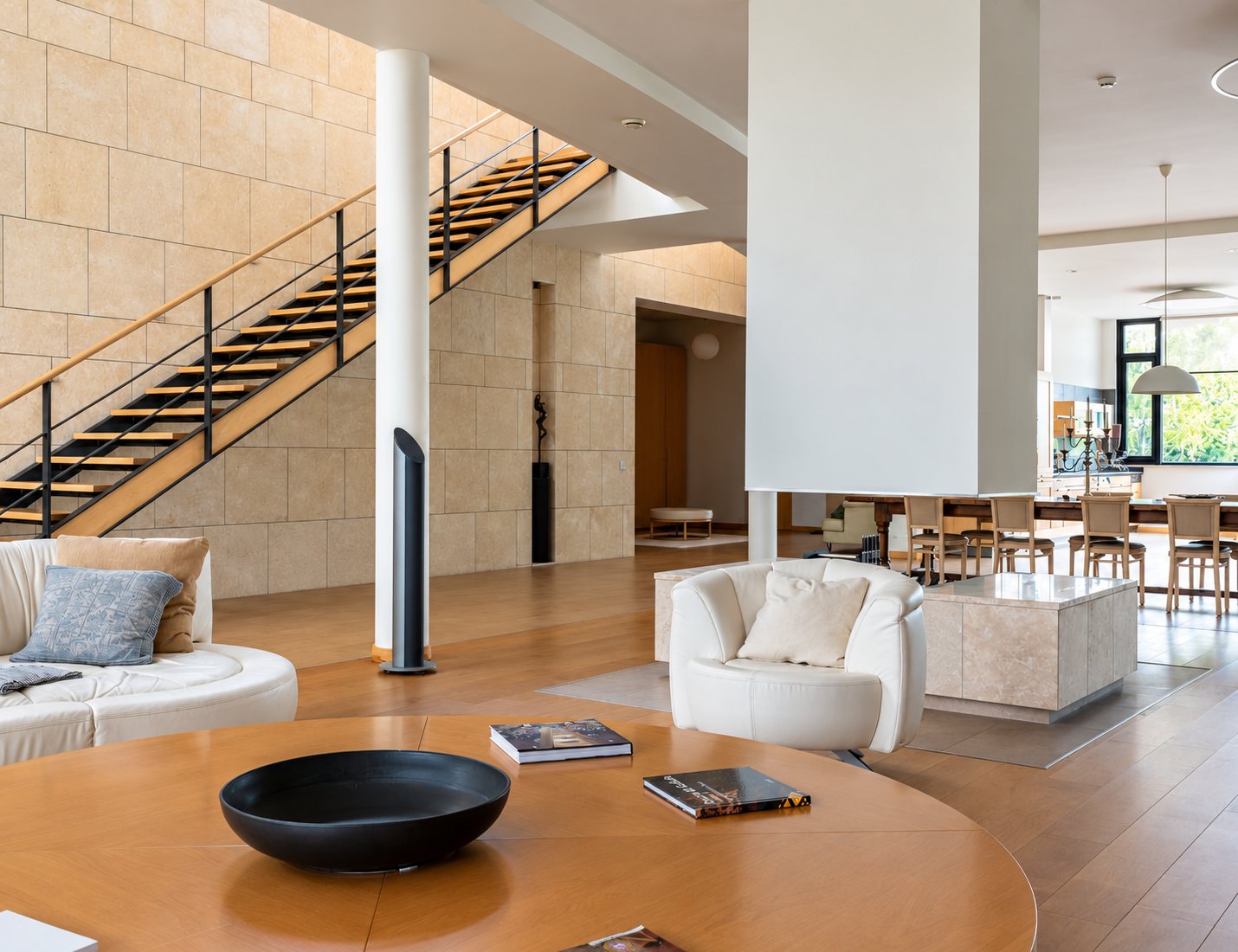 Vaste salon lumineux au style contemporain, avec un escalier à marches en bois, un fauteuil blanc, une table ronde en bois et une salle à manger en arrière-plan.