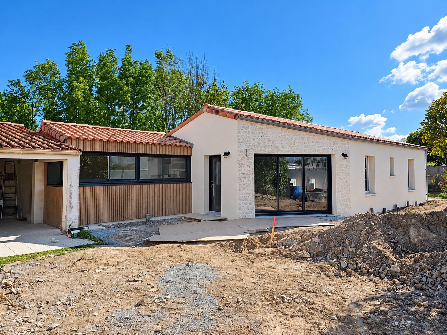 Maison récente de plain-pied aux murs crépis, pierre et bardage bois, avec baie vitrée noire et terrassement en cours au premier plan.