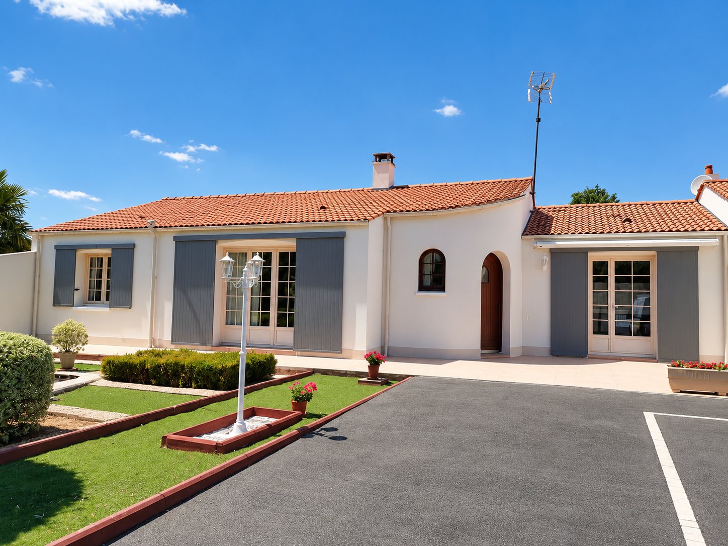 Maison de plain-pied aux murs clairs et au toit de tuiles, avec volets gris, porte-fenêtres, pelouse fleurie et allée goudronnée sous un ciel bleu.