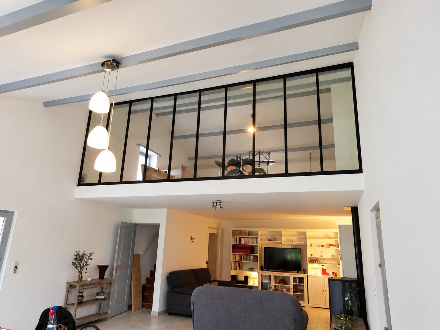 Une mezzanine fermée par une grande verrière à cadres noirs surplombe un salon avec canapés, bibliothèque blanche et suspension à trois globes.