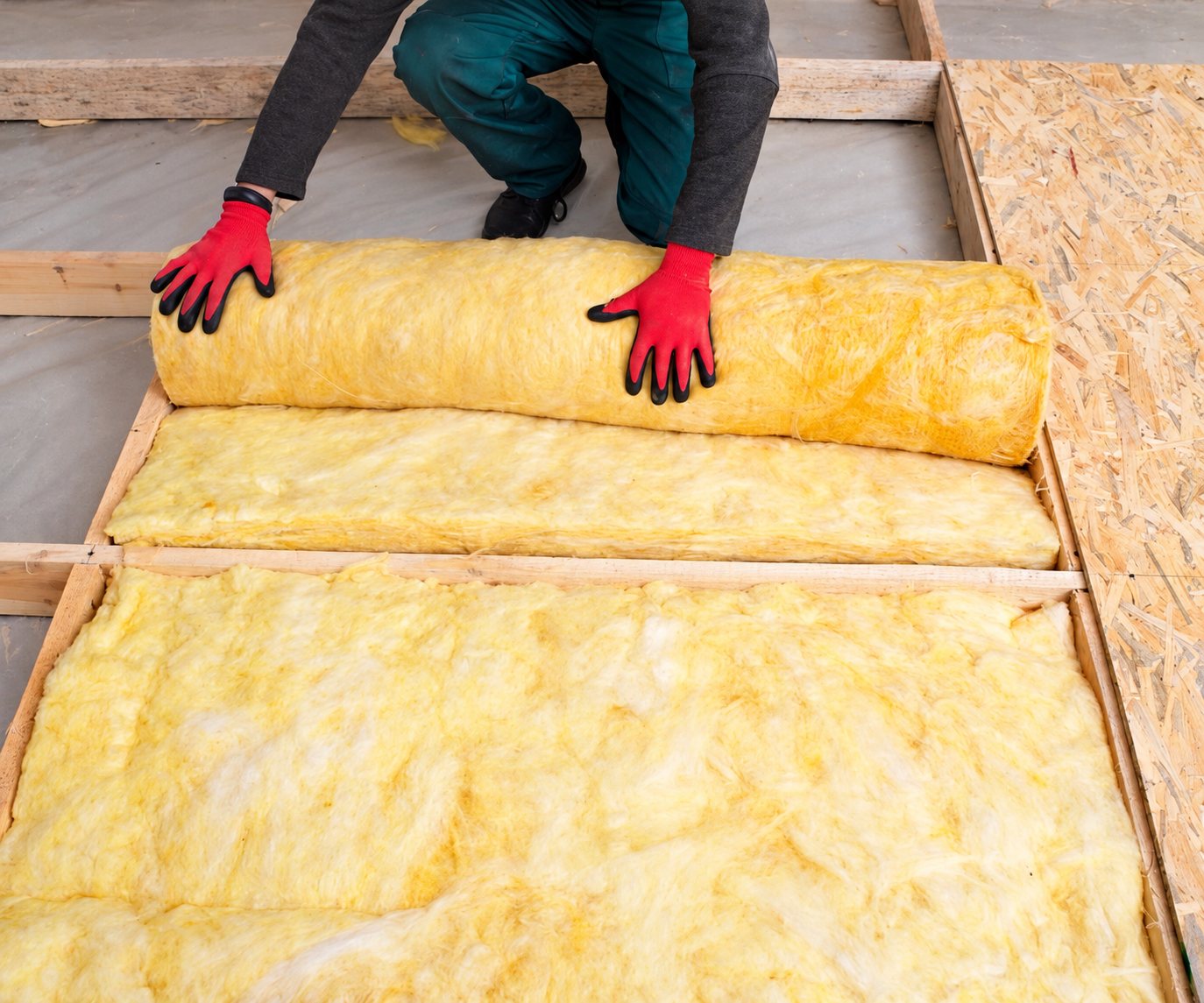 Un ouvrier aux gants rouges déroule un rouleau de laine de verre jaune entre des solives en bois sur un plancher en panneaux OSB.