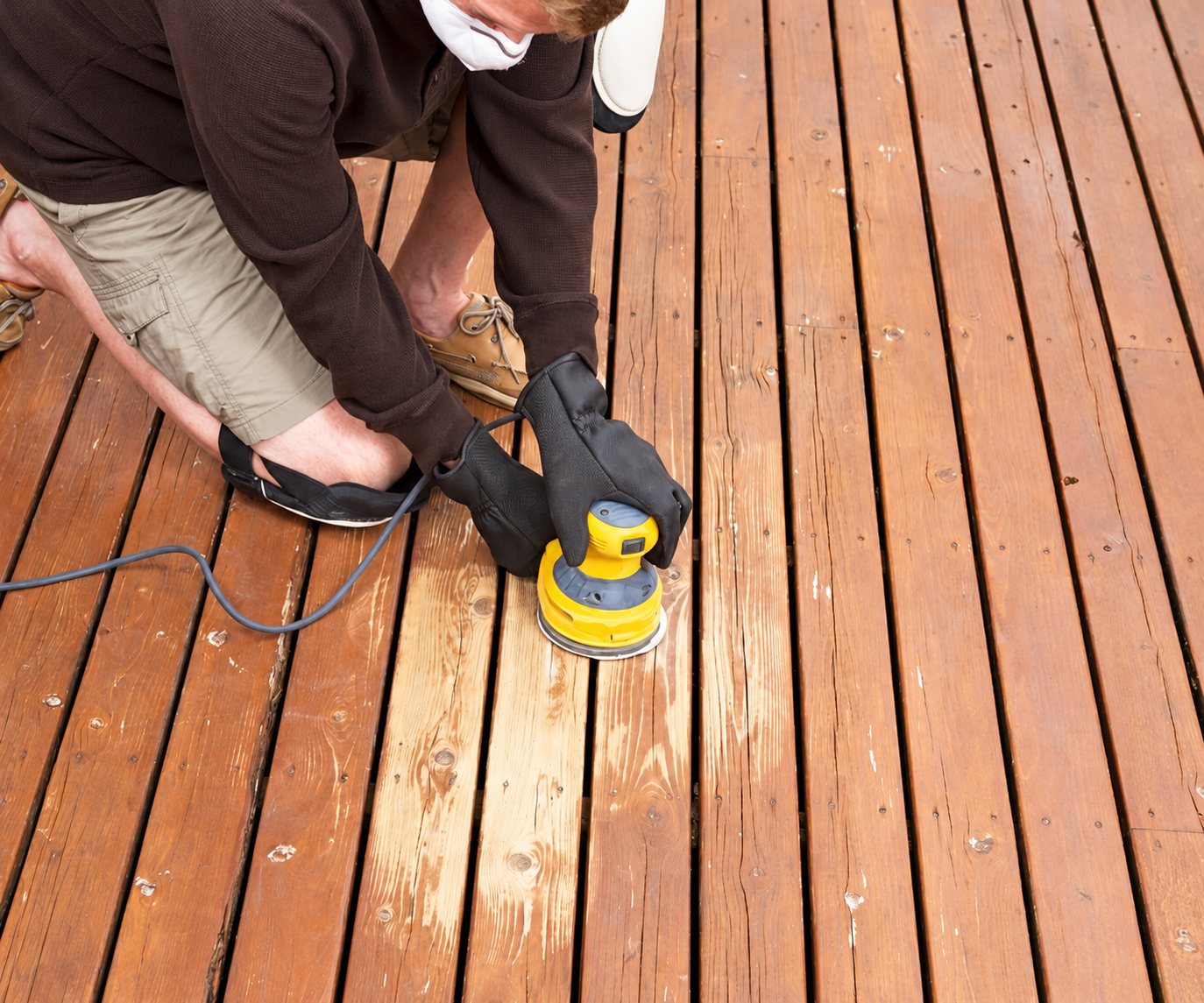Un homme agenouillé sur une terrasse en bois ponce les lames avec une ponceuse orbitale jaune, portant des gants et un masque.