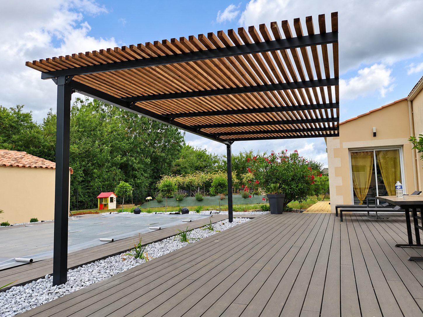 Une pergola à structure métallique sombre et lames de bois couvre une terrasse en lames grises, à côté d'une piscine bâchée et d'un massif de galets blancs.