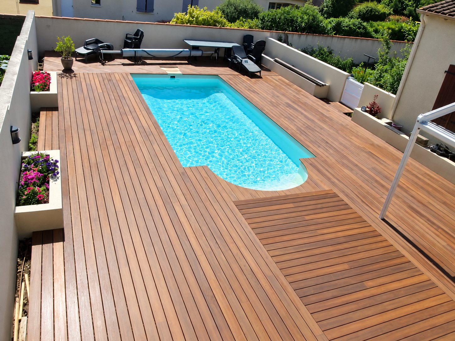 Vue en hauteur d'une piscine rectangulaire à l'eau turquoise entourée d'une terrasse en lames de bois, de jardinières fleuries et de murs blancs.