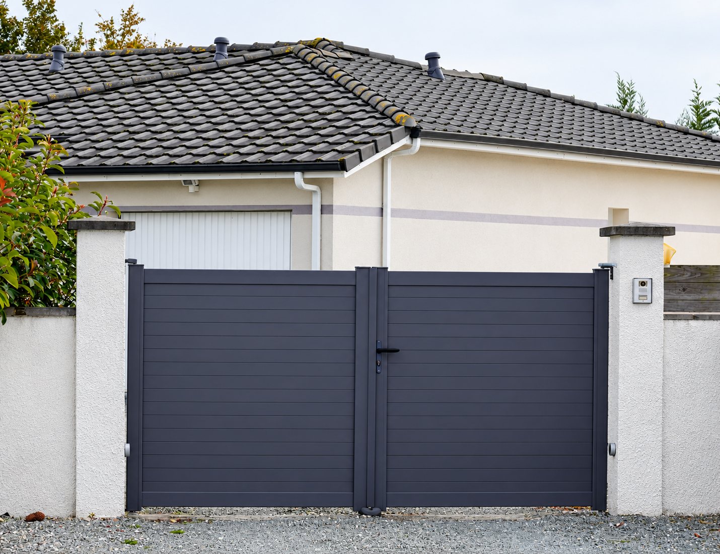 Un portail battant à deux vantaux en aluminium gris anthracite, entre deux piliers blancs, devant une maison au toit de tuiles grises.