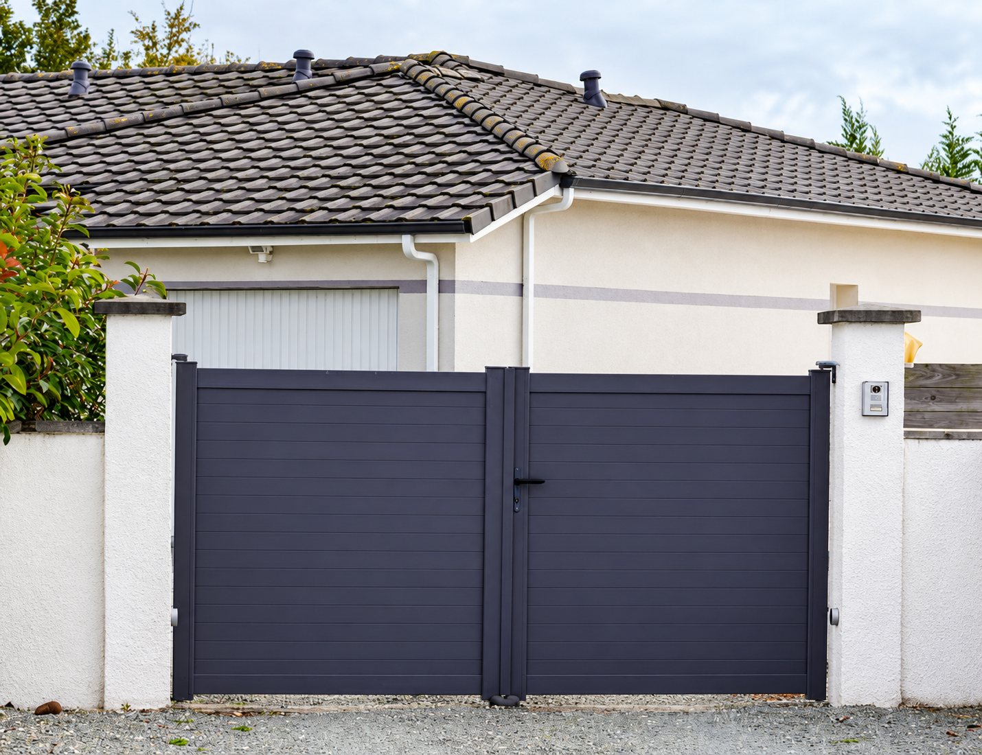 Un portail battant à deux vantaux en aluminium gris anthracite est installé entre deux piliers blancs devant une maison à toit de tuiles grises.