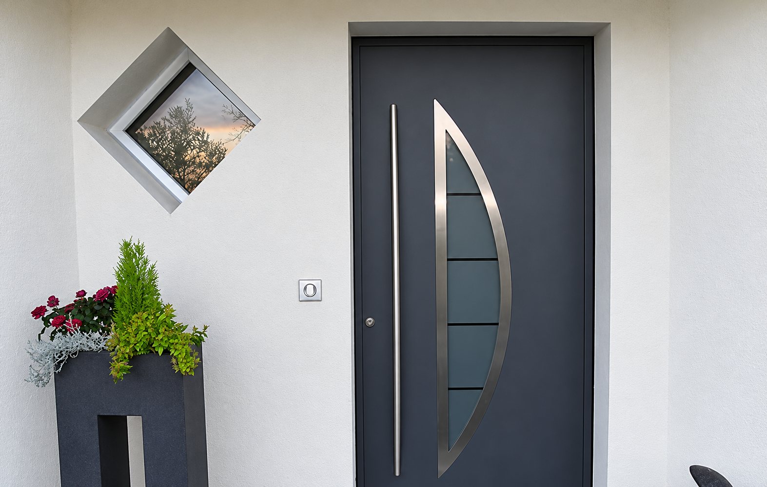 Porte d'entrée gris anthracite avec longue poignée en inox et vitrage en demi-lune, installée dans une façade blanche à côté d'une fenêtre en losange et d'une jardinière fleurie.
