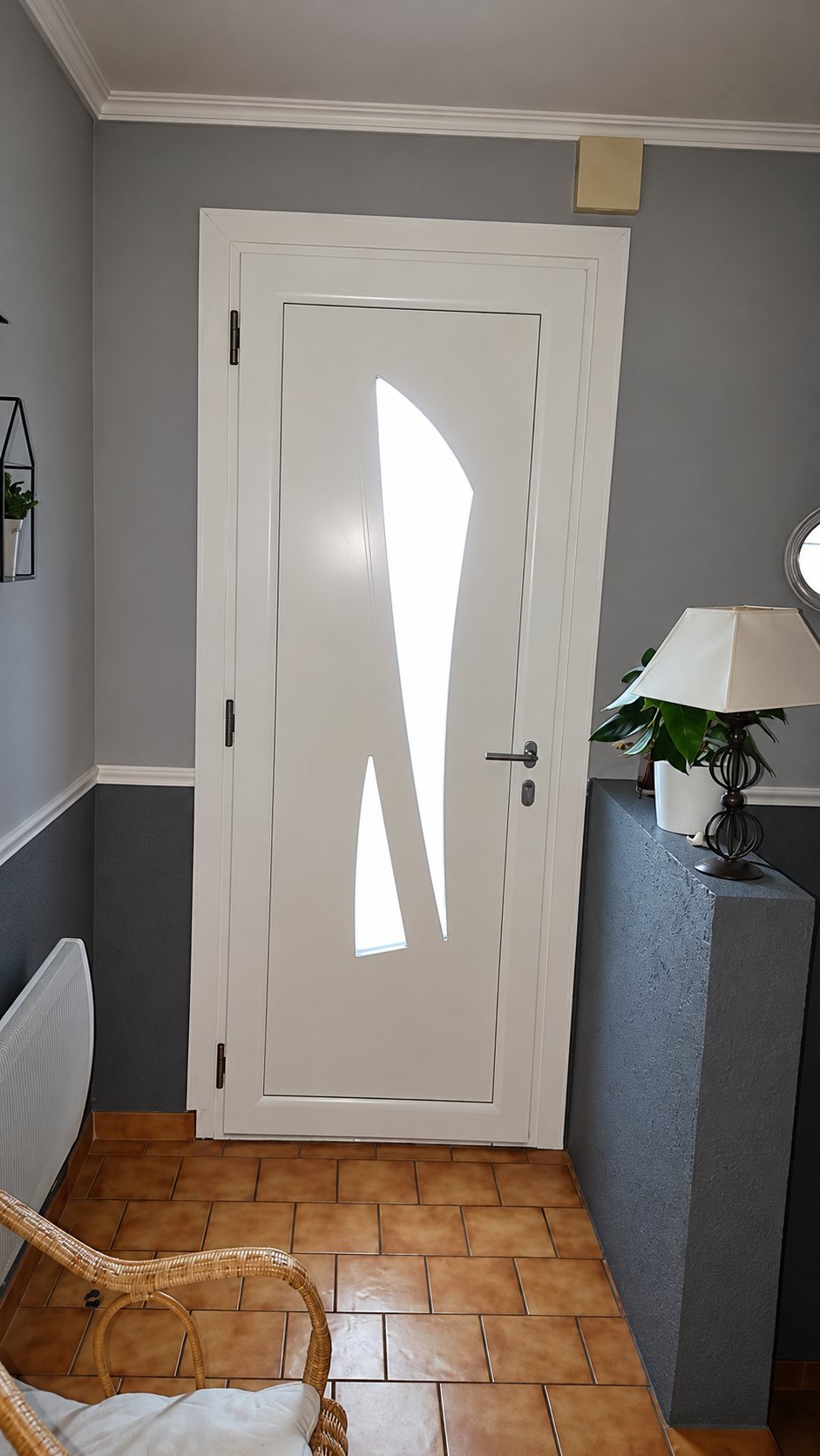 Une porte d'entrée blanche avec une fenêtre étroite aux formes courbes, installée dans un hall aux murs gris et au carrelage orangé, près d'une lampe et d'une plante.