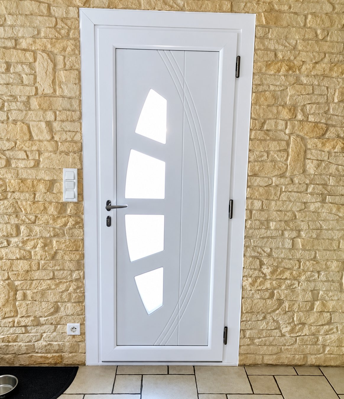 Porte d'entrée blanche avec quatre petits vitrages en arc et rainures décoratives, posée dans un mur de pierres claires au-dessus d'un carrelage.