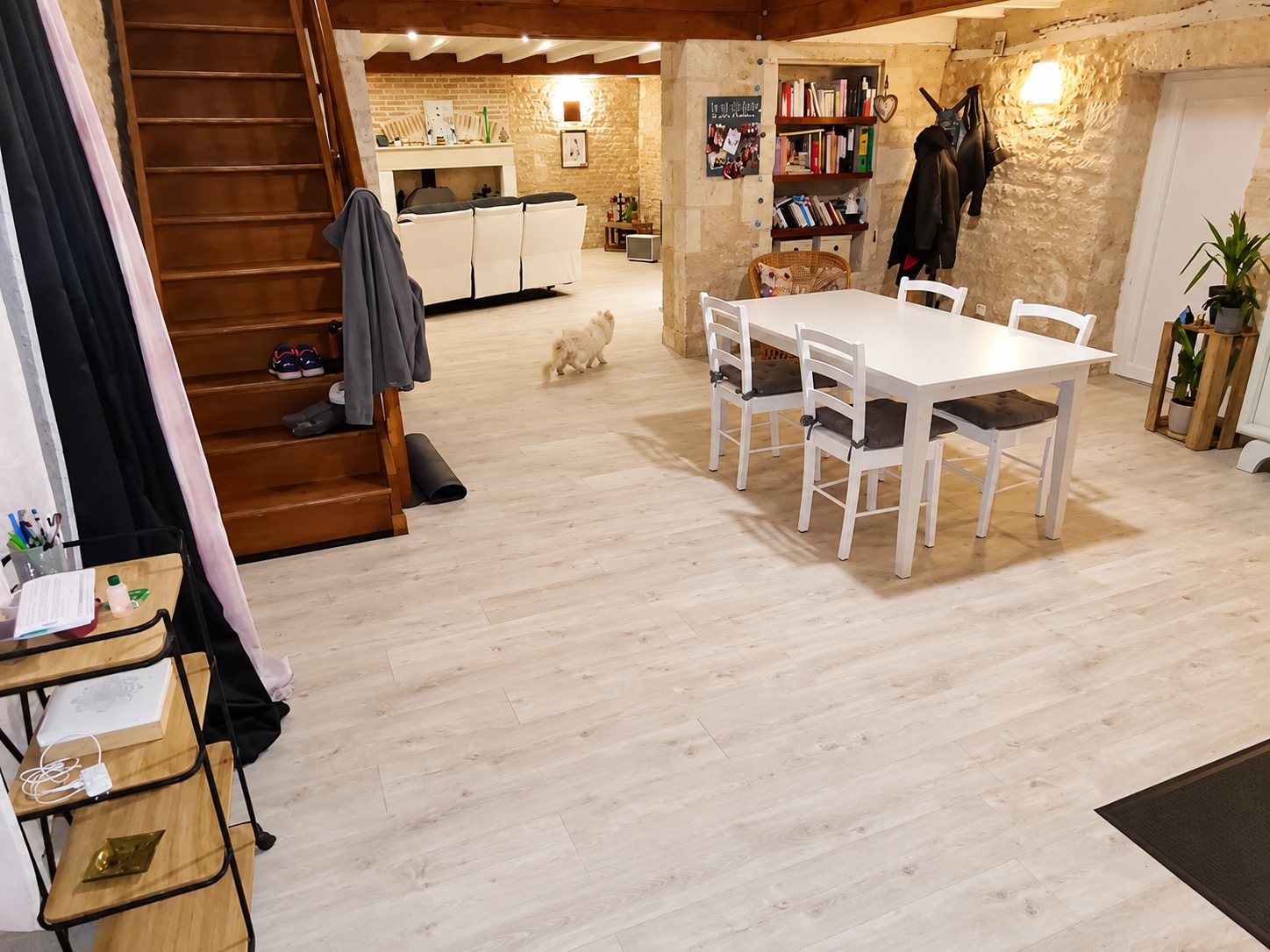Grande pièce de vie aux murs en pierre apparente avec un sol clair neuf, une table blanche entourée de chaises, un escalier en bois et un petit chien blanc.