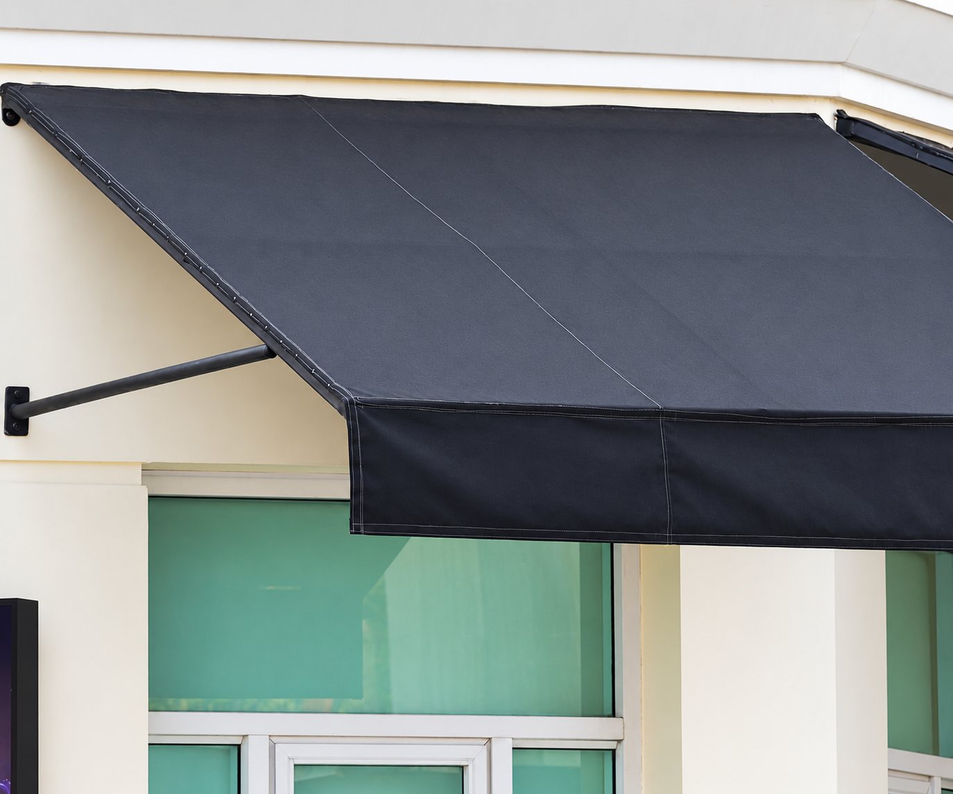 Un store fixe en toile gris anthracite, soutenu par un bras métallique, est installé sur une façade crème au-dessus de grandes fenêtres vitrées.