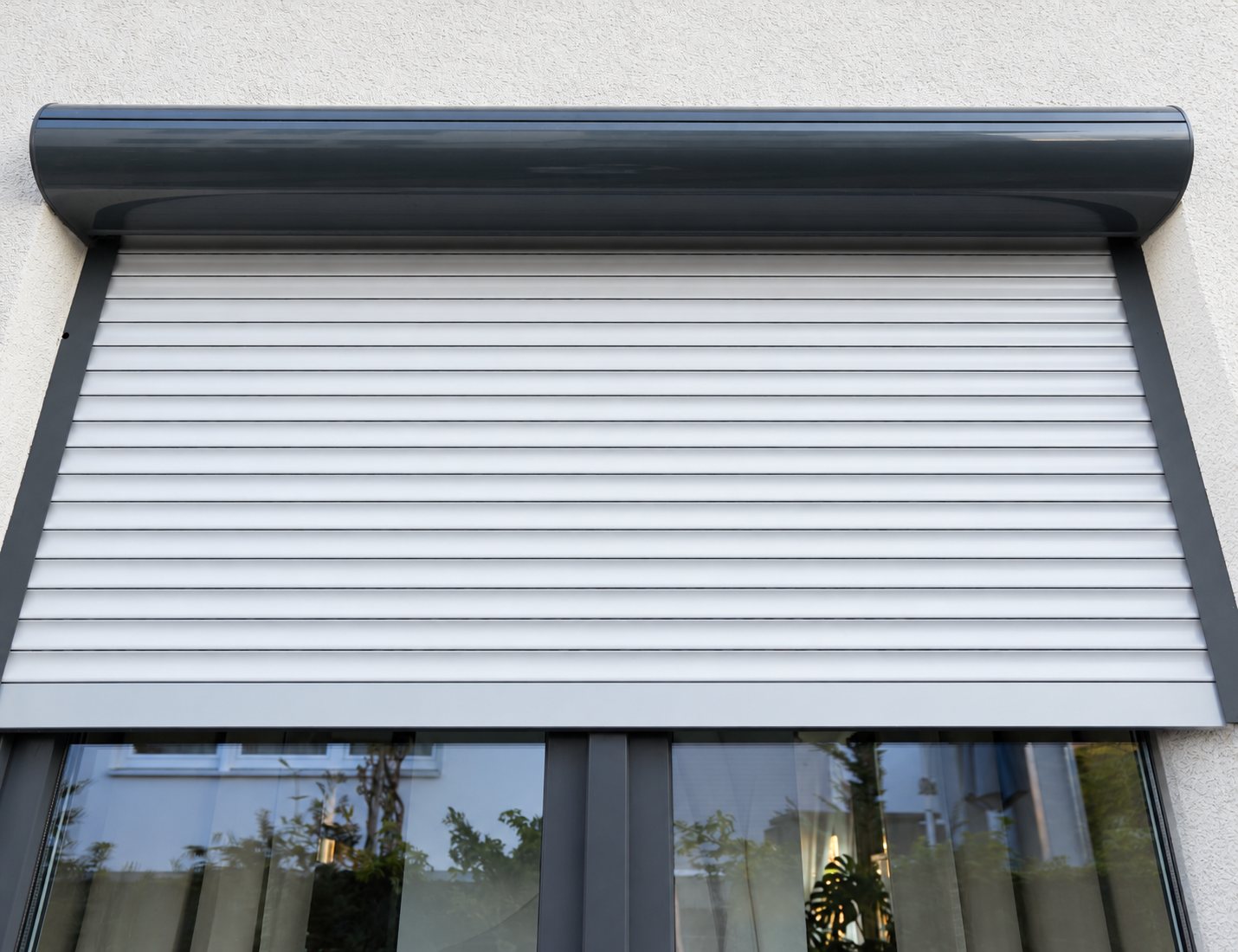Un volet roulant en aluminium blanc, avec son caisson arrondi gris anthracite, est posé au-dessus d'une fenêtre sur un mur crépi clair.