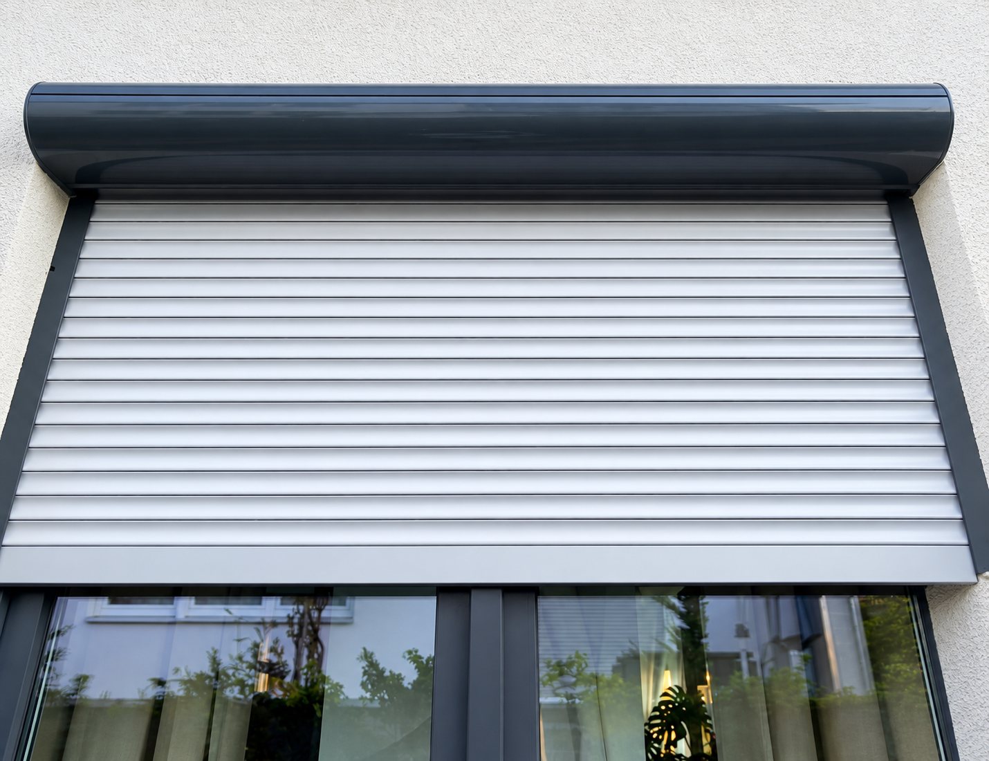 Un volet roulant en lames blanches, avec son caisson gris anthracite, est posé au-dessus d'une grande fenêtre sur un mur blanc.